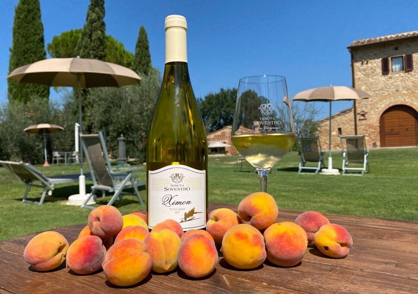 A bottle of Tenuta Sovestro wine, a glass of wine, and several peaches on a wooden table outdoors.