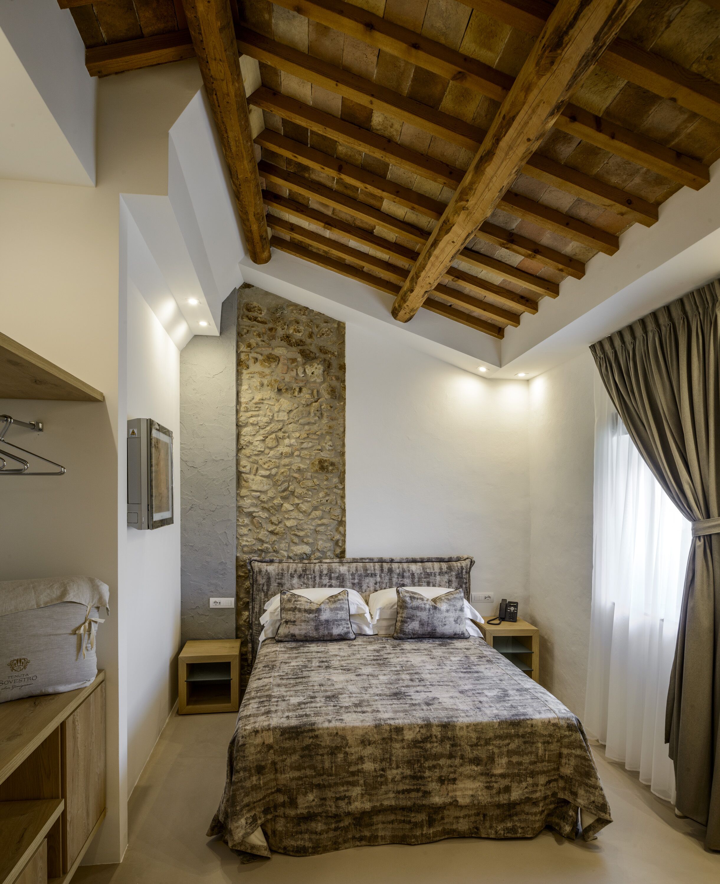 A bedroom with a stone wall, wooden ceiling beams, a bed with a textured blanket, a bedside table with a lamp, and a window with curtains.