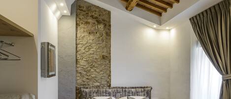 A bedroom with a stone wall, wooden ceiling beams, a bed with a textured blanket, a bedside table with a lamp, and a window with curtains.