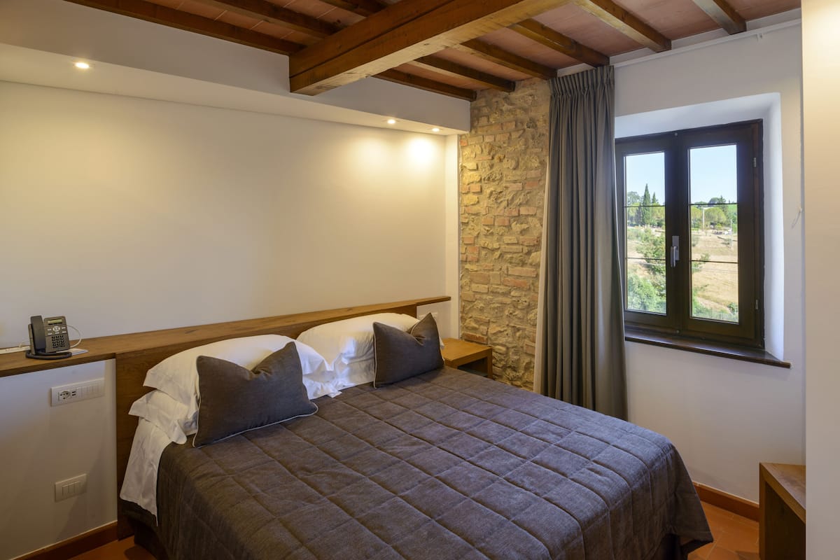 A bedroom with a bed, a stone wall, a window with curtains, and a wooden ceiling.