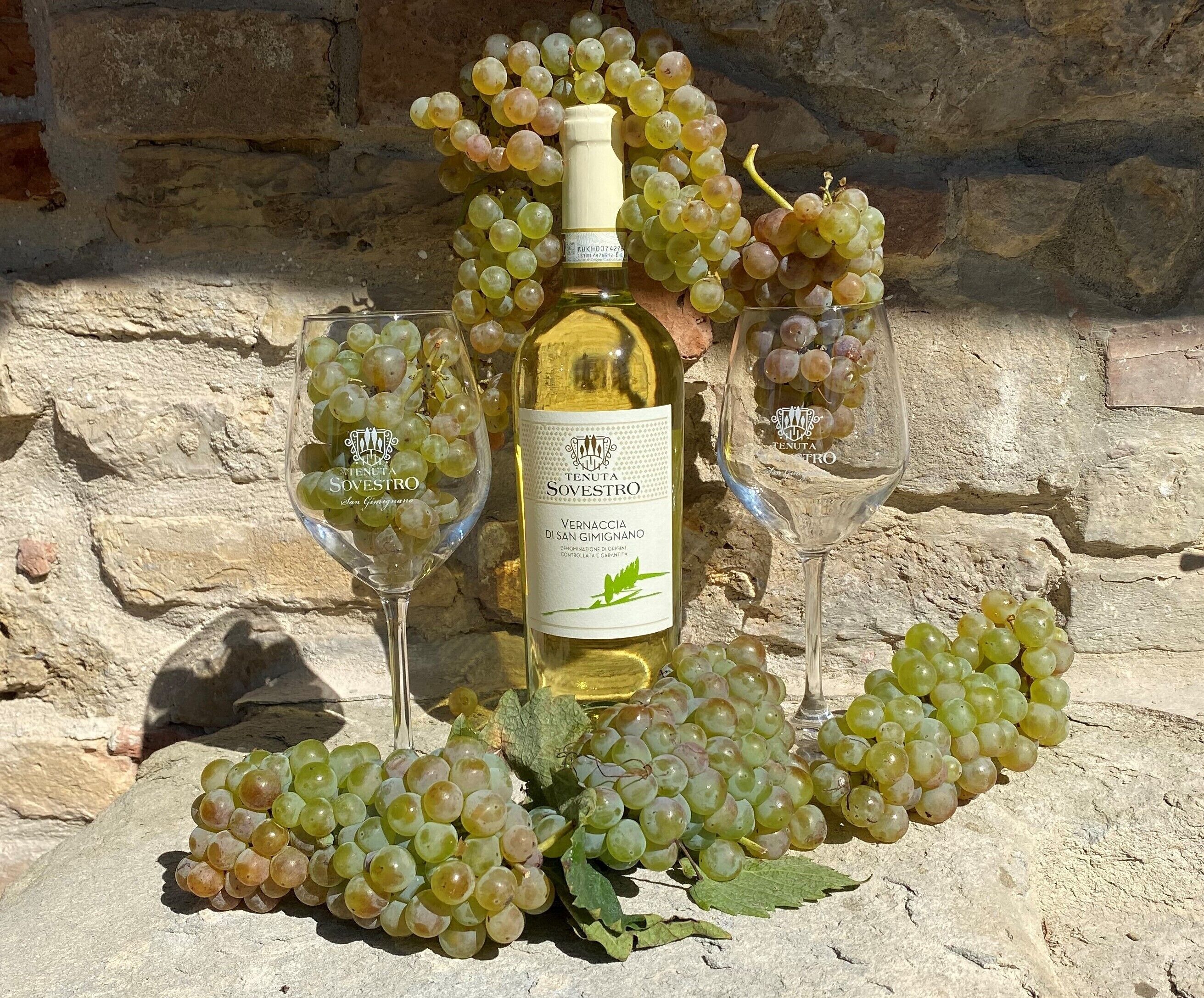Una bottiglia di vino con etichetta, due bicchieri e grappoli d'uva su un muro in pietra.
