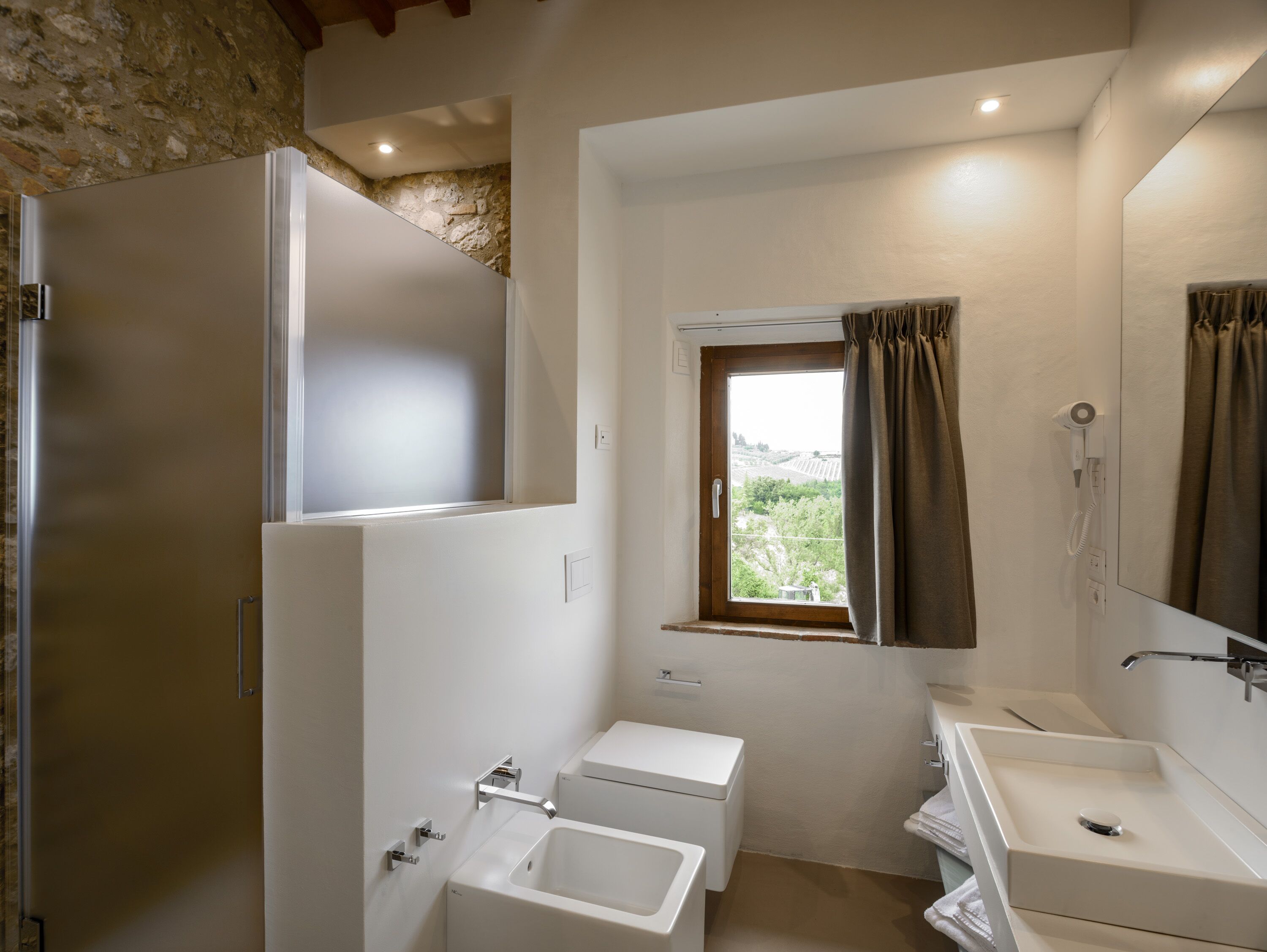 A bathroom with a shower, toilet, and sink, a window with curtains, and a stone wall.