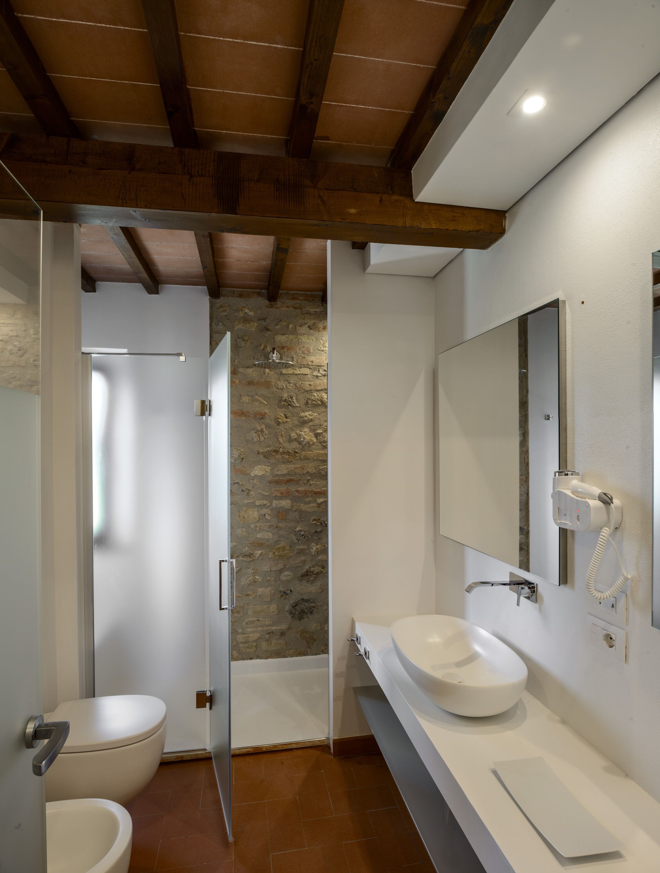 Une salle de bain avec un mur en pierre, un lavabo blanc, un miroir et une douche.