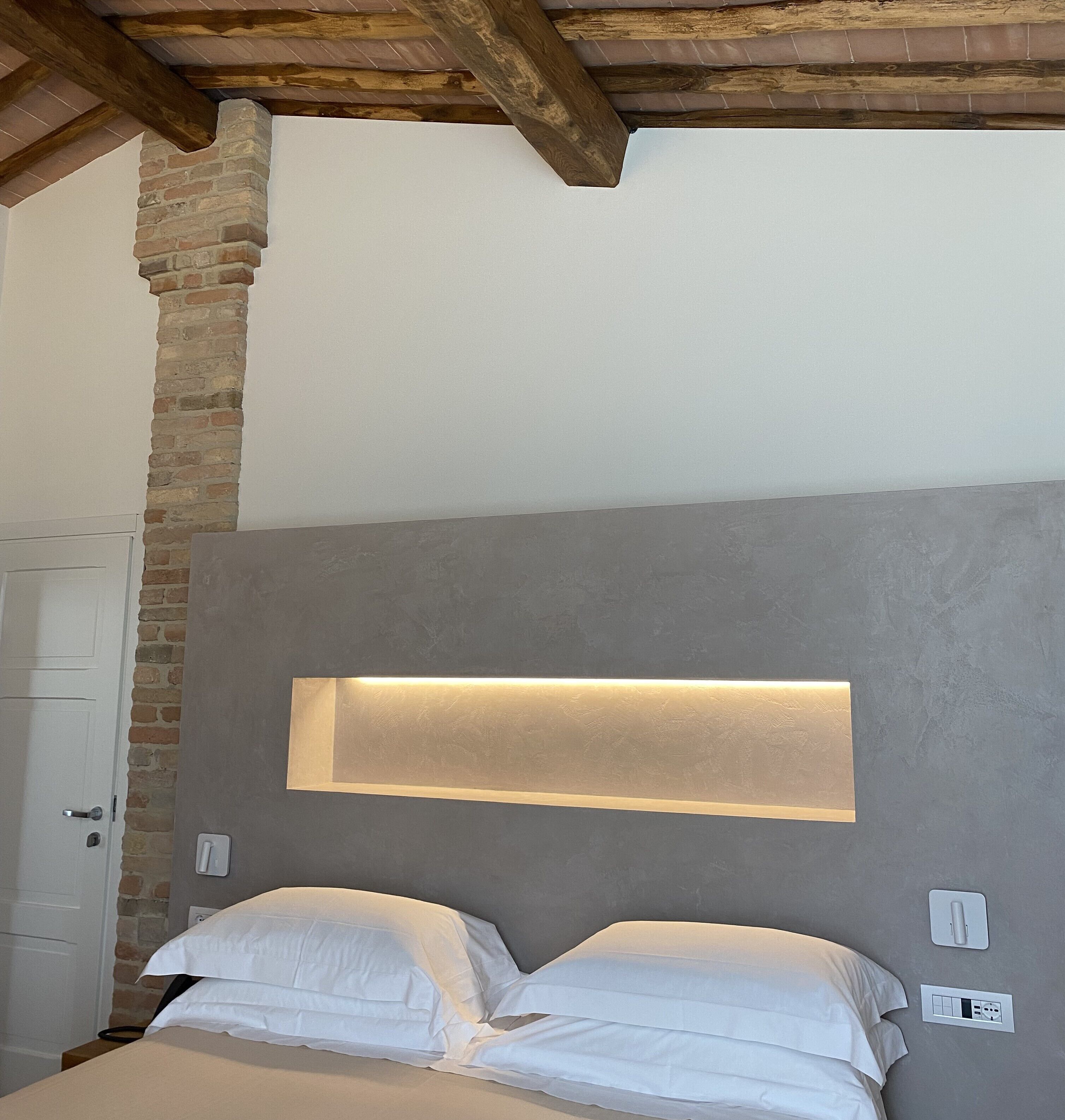 A bedroom with a bed, a wall-mounted light, a brick column, and a door.