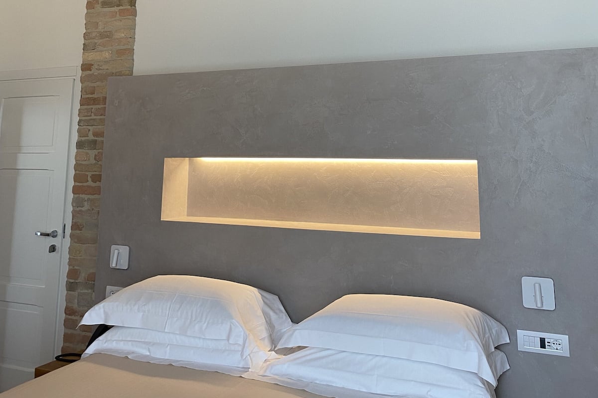 A bedroom with a bed, a wall-mounted light, a brick column, and a door.