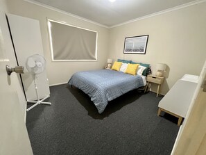 A bedroom with a bed, bedside tables, a standing fan, a window with blinds, and a framed picture on the wall. - Caboolture, Central Location, Sleeps 6 (Caboolture South)