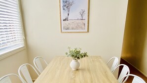 A dining area with a wooden table, white chairs, a vase with greenery, and a framed picture on the wall. - Caboolture, Central Location, Sleeps 6 (Caboolture South)