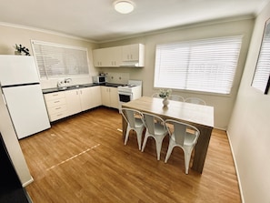 A compact kitchen with white cabinets, a wooden countertop, a dining table with four chairs, and a window with blinds. - Caboolture, Central Location, Sleeps 6 (Caboolture South)
