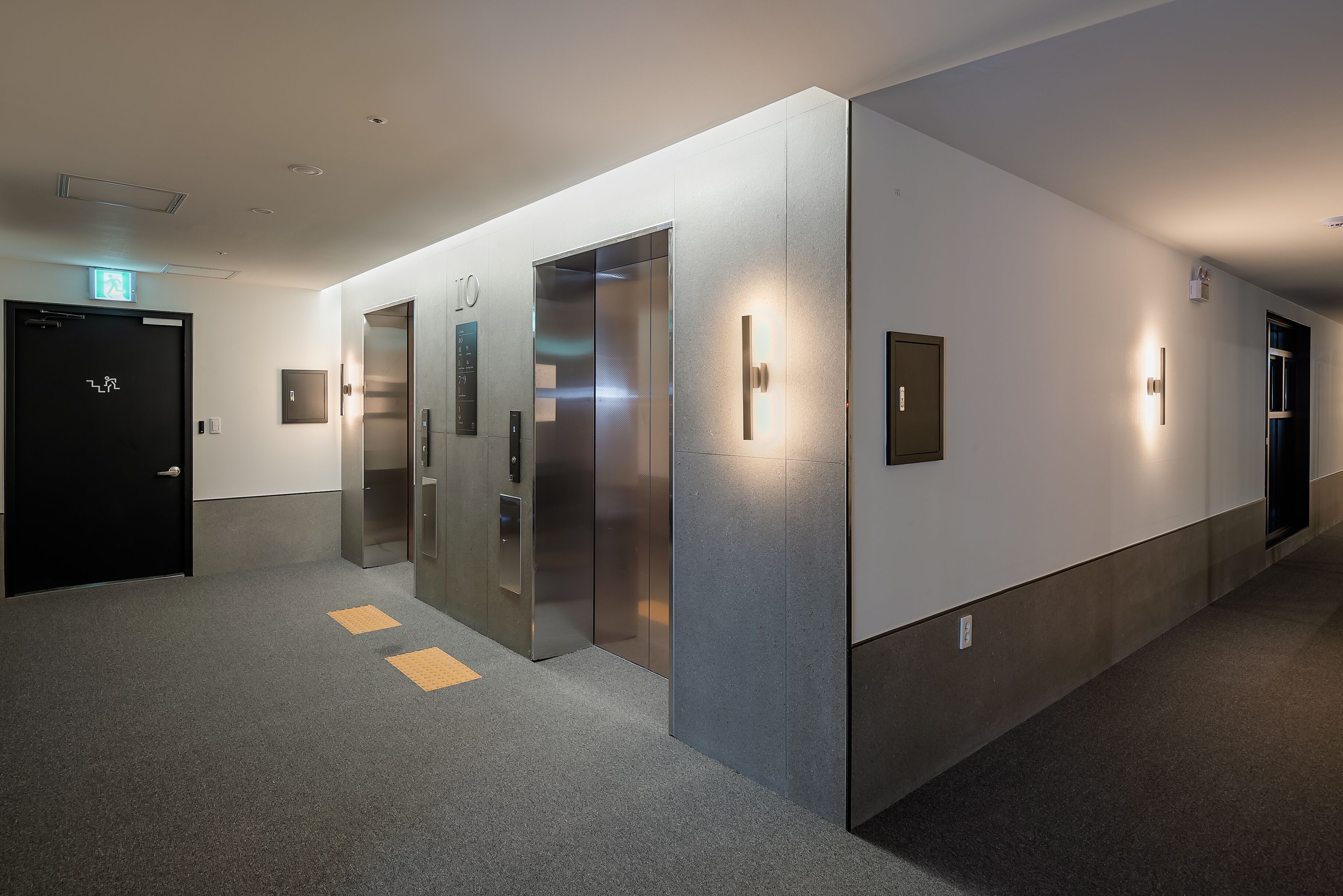 Un pasillo moderno con tres ascensores, una puerta negra con letrero de salida y un elemento decorativo iluminado en la pared.