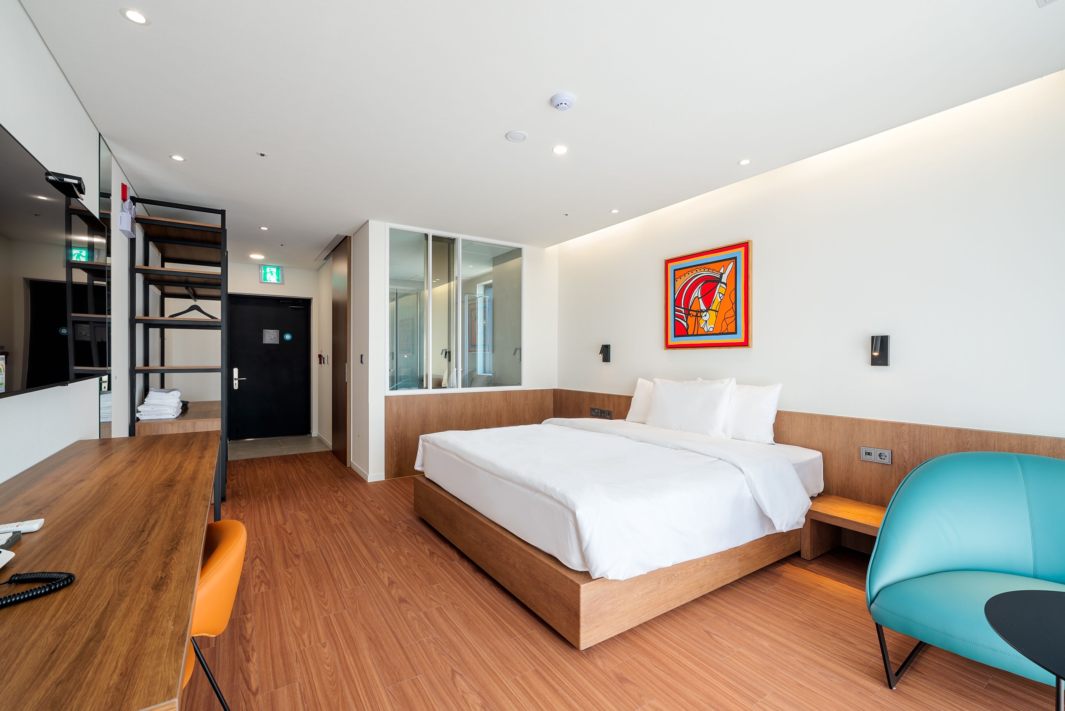 A modern hotel room with a wooden floor, a large bed with white linens, a blue armchair, a desk with a telephone, and a colorful artwork on the wall.