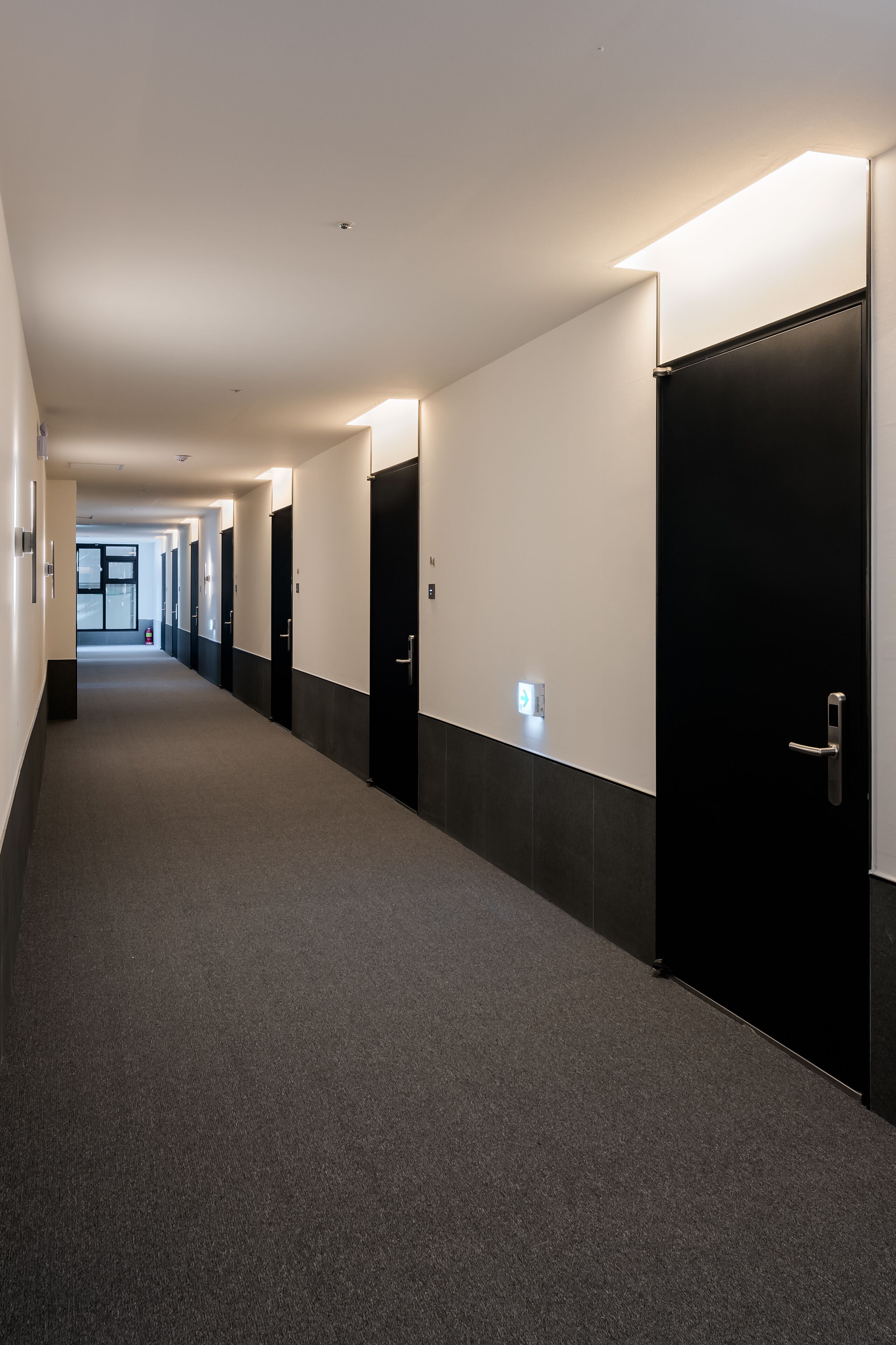 Un long couloir étroit aux murs noirs et blancs, avec des portes de chaque côté et un sol recouvert de moquette.
