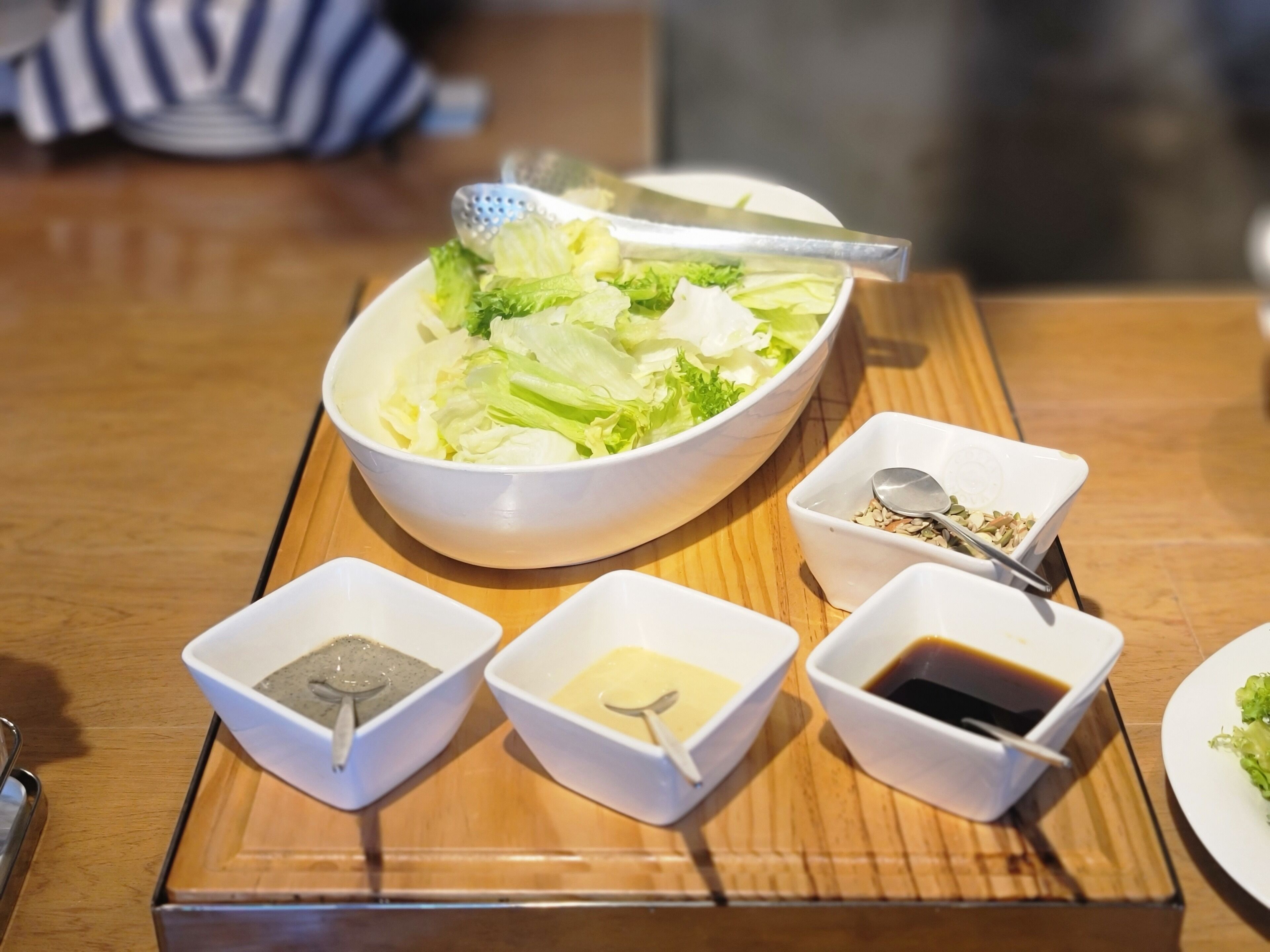 A wooden table with a white bowl of salad, three small bowls with condiments, and a spoon in one of the bowls.