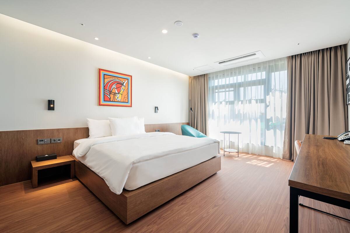A modern hotel room with a large bed, a desk, and a chair. There is a painting on the wall and a window with curtains.