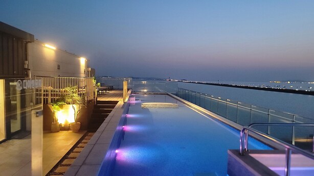 A rooftop pool area with a view of the sea, a glass railing, and a seating area with plants. - Hotel Cielo (Siheung)