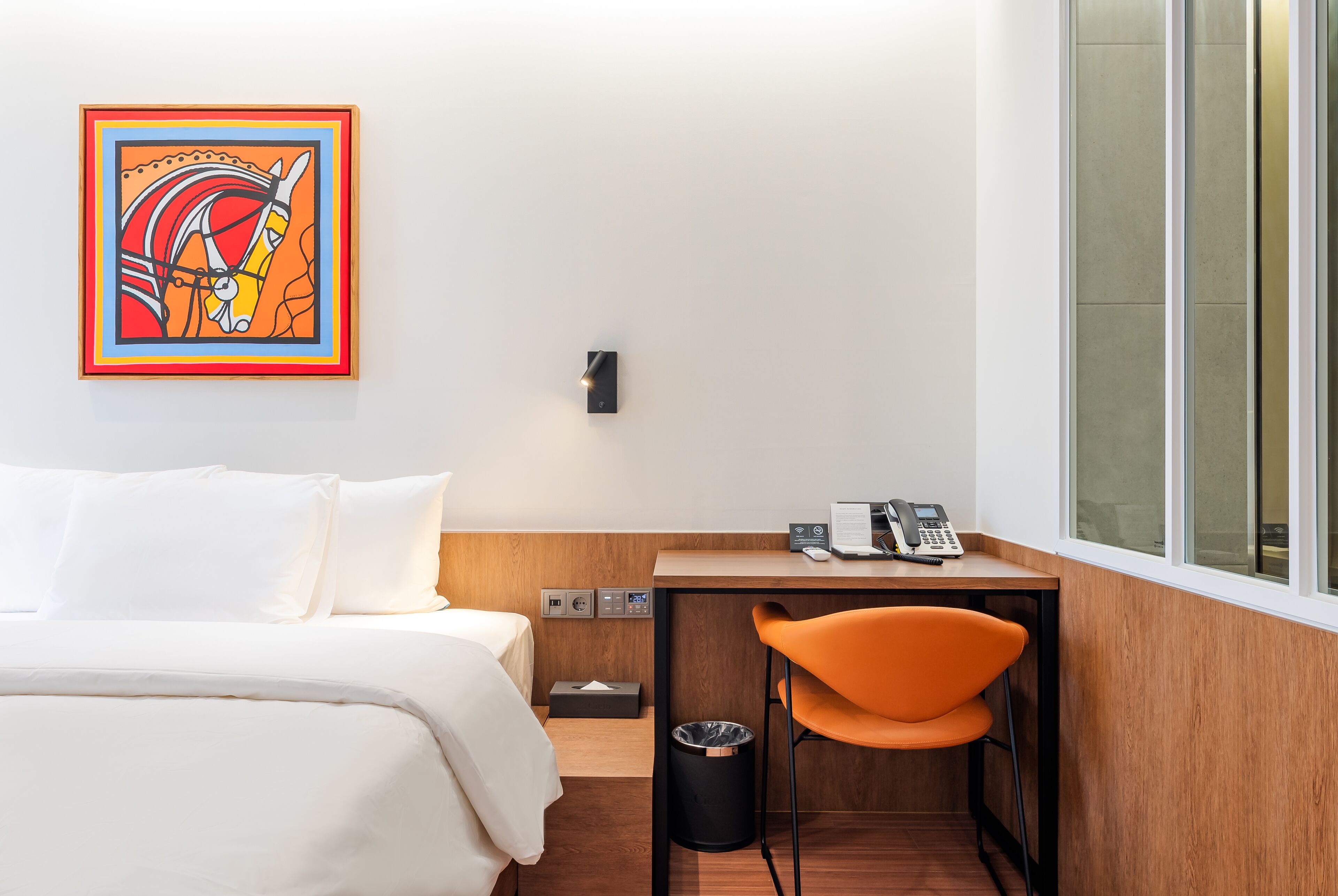 A hotel room with a bed, a desk with a chair, a telephone, a trash bin, and a wall-mounted light.