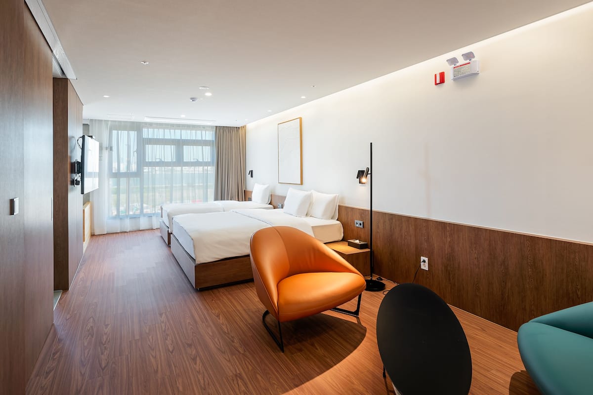 A modern hotel room with a large bed, a wooden floor, a desk with a chair, and a large window with curtains.