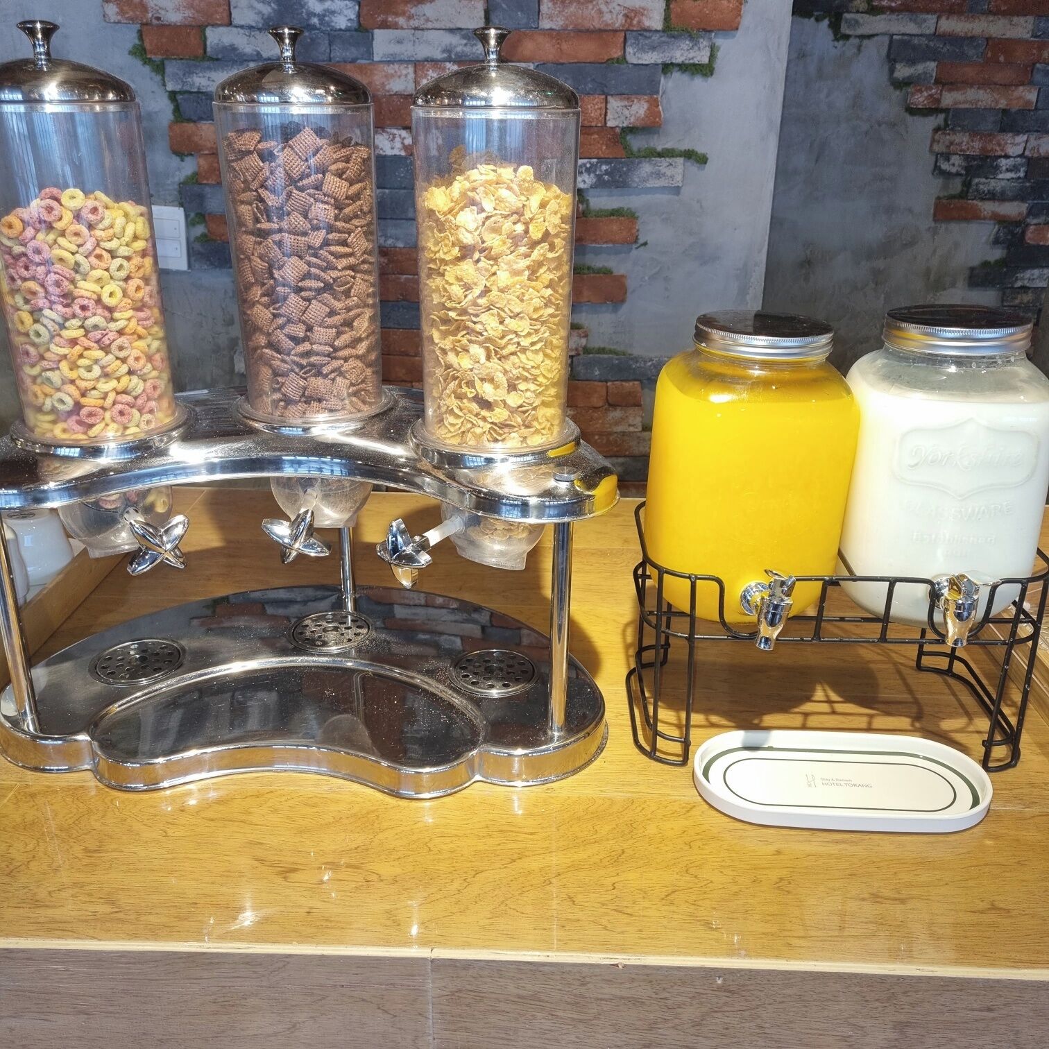 Una estación de bebidas autoservicio con tres dispensadores para distintos tipos de cereales y dos grandes recipientes para jugo de naranja y leche.
