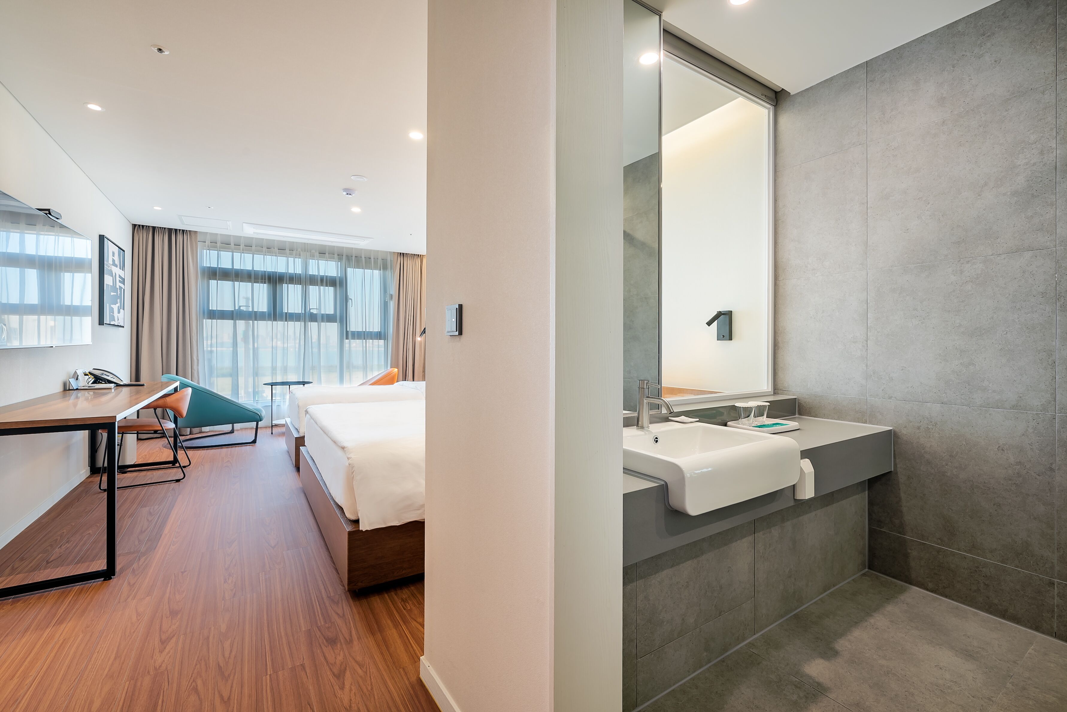 A modern hotel room with a wooden floor, a bed with white linens, a desk with a chair, and a large mirror above the sink.