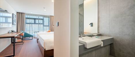 A modern hotel room with a wooden floor, a bed with white linens, a desk with a chair, and a large mirror above the sink.