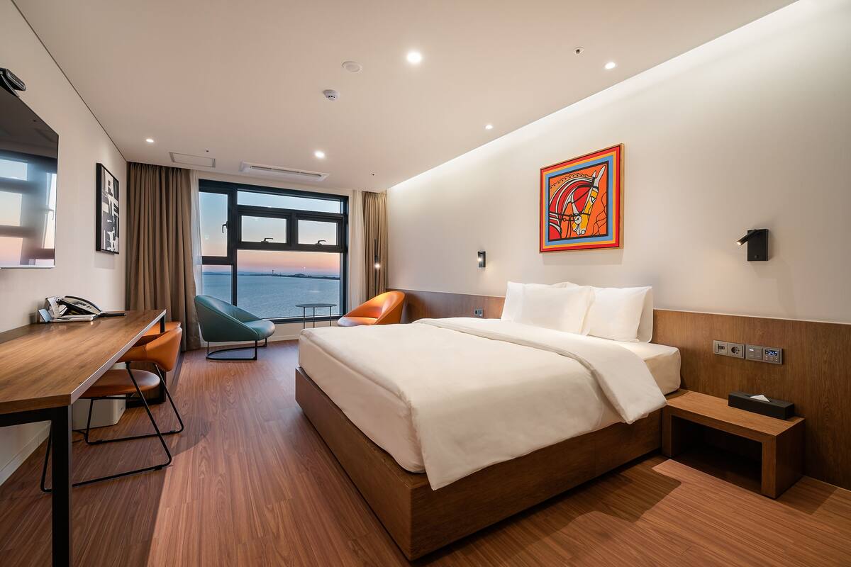 A modern hotel room with a large bed, a desk, a chair, and a view of the sea.