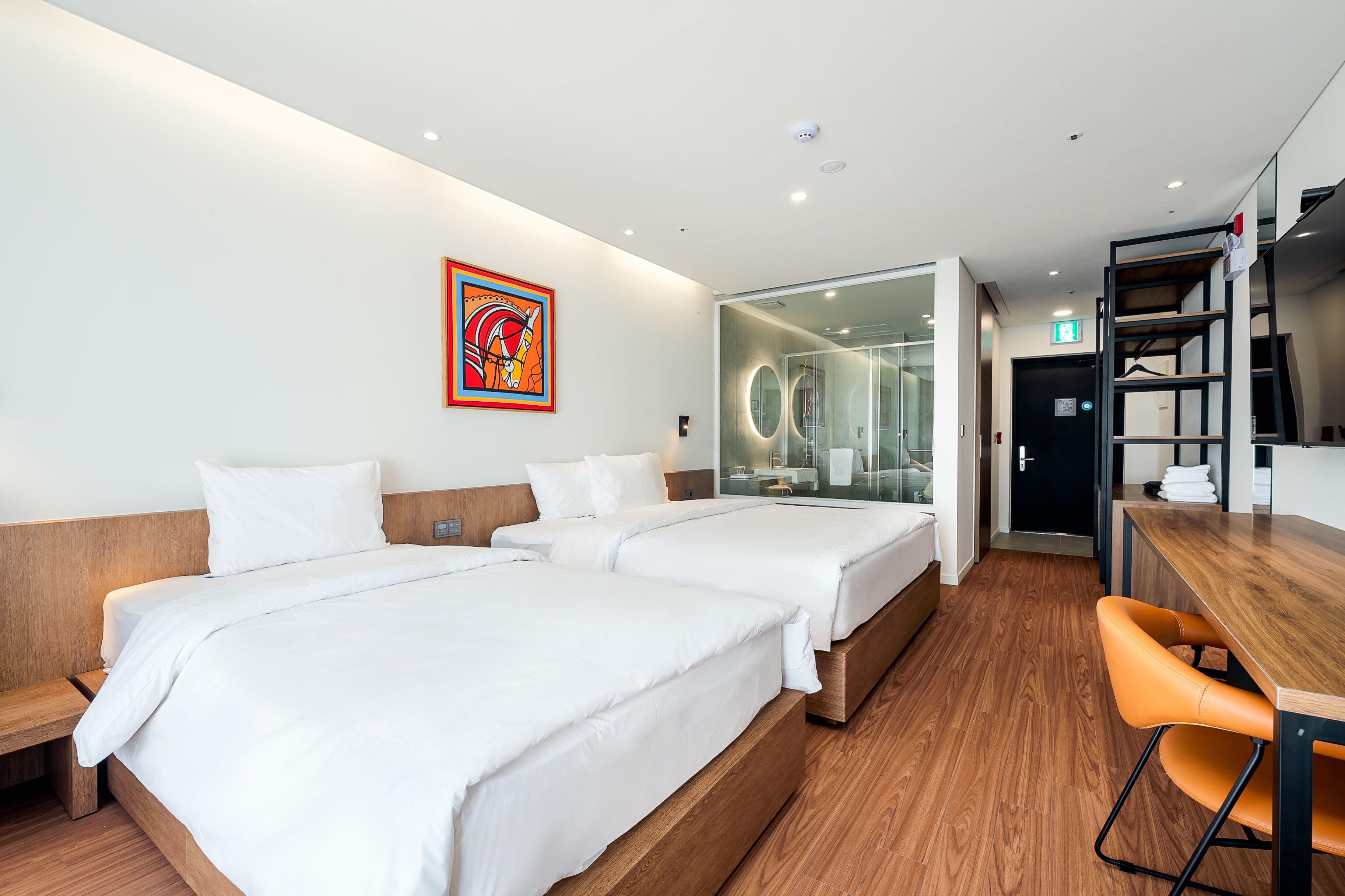 Habitación de hotel con dos camas, suelo de madera, escritorio con silla y baño visible a través de una puerta de cristal.