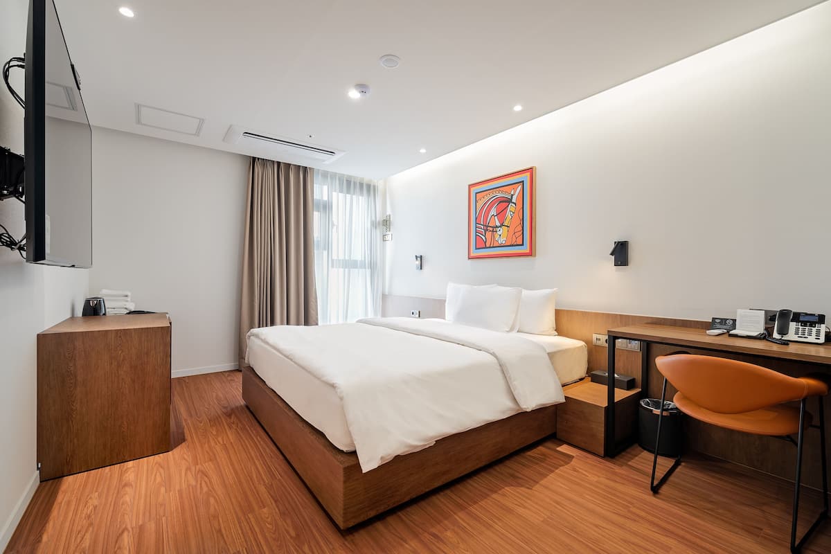 A modern hotel room with a wooden bed, a desk with a chair, a flat-screen TV, and a painting on the wall.