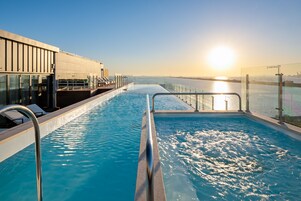 Zona de piscina con barandilla de cristal, sillas de descanso y vista al atardecer sobre el agua.