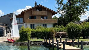 A wooden house with a balcony, a sailboat docked at a wooden pier, and a stone wall in the background. - Garden Suite with cozy eat-in kitchen and lake view (Frauenchiemsee)