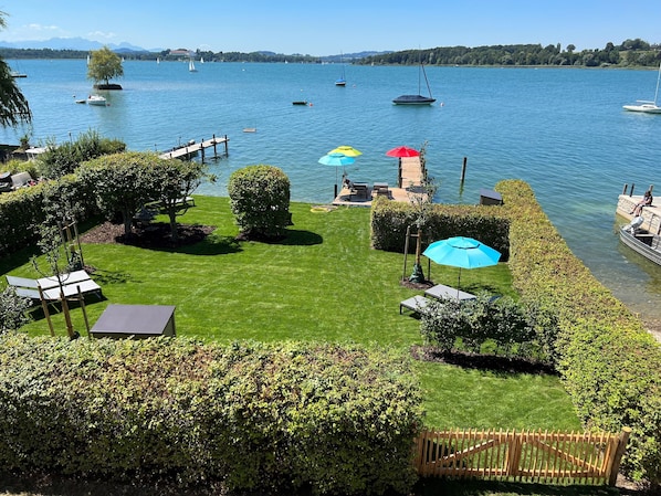 A well-maintained garden with a wooden deck overlooking a lake, featuring a lawn, several umbrellas, and a wooden fence.