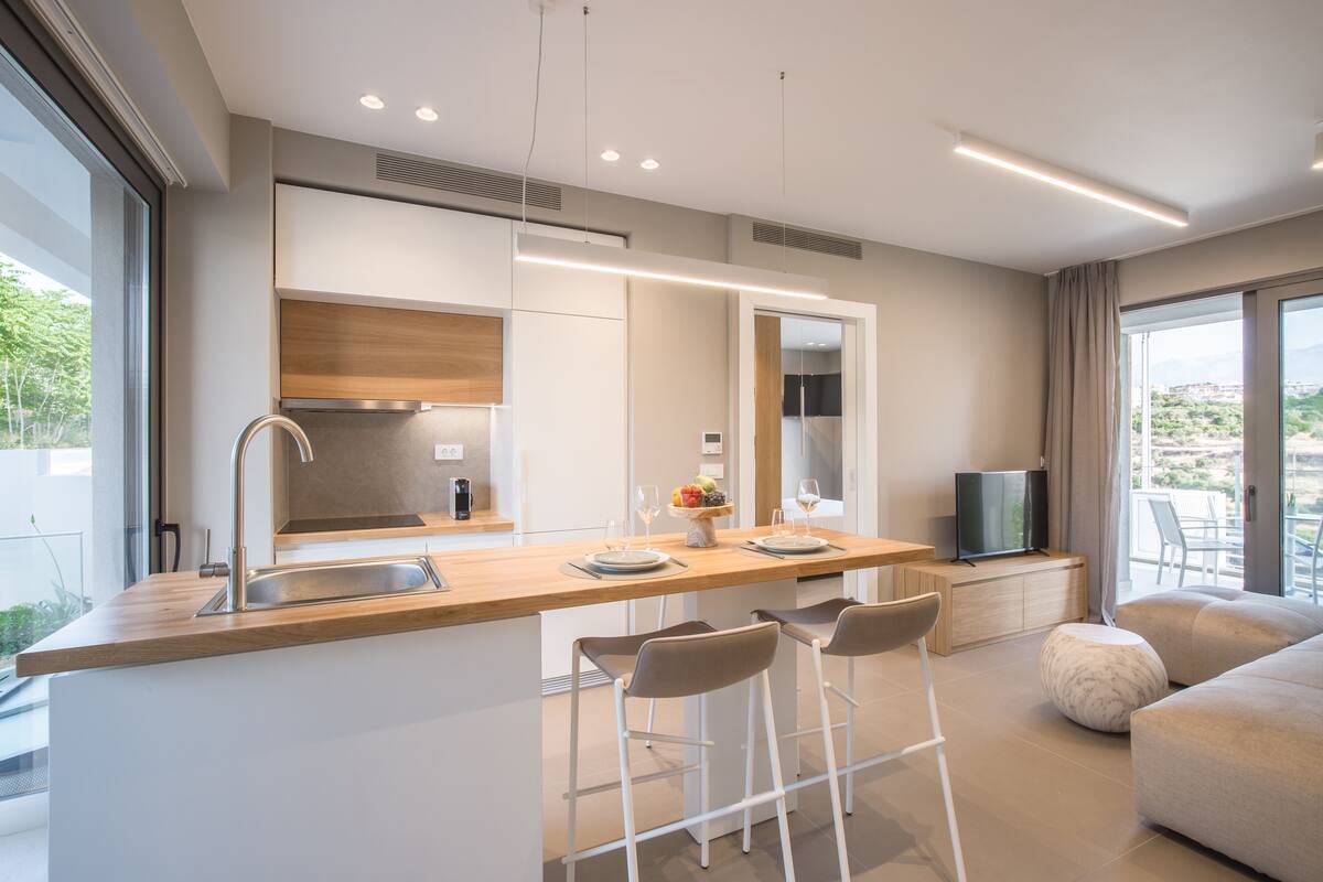 Una cocina moderna con una isla central, taburetes de bar, horno empotrado y un televisor sobre un mueble de madera.