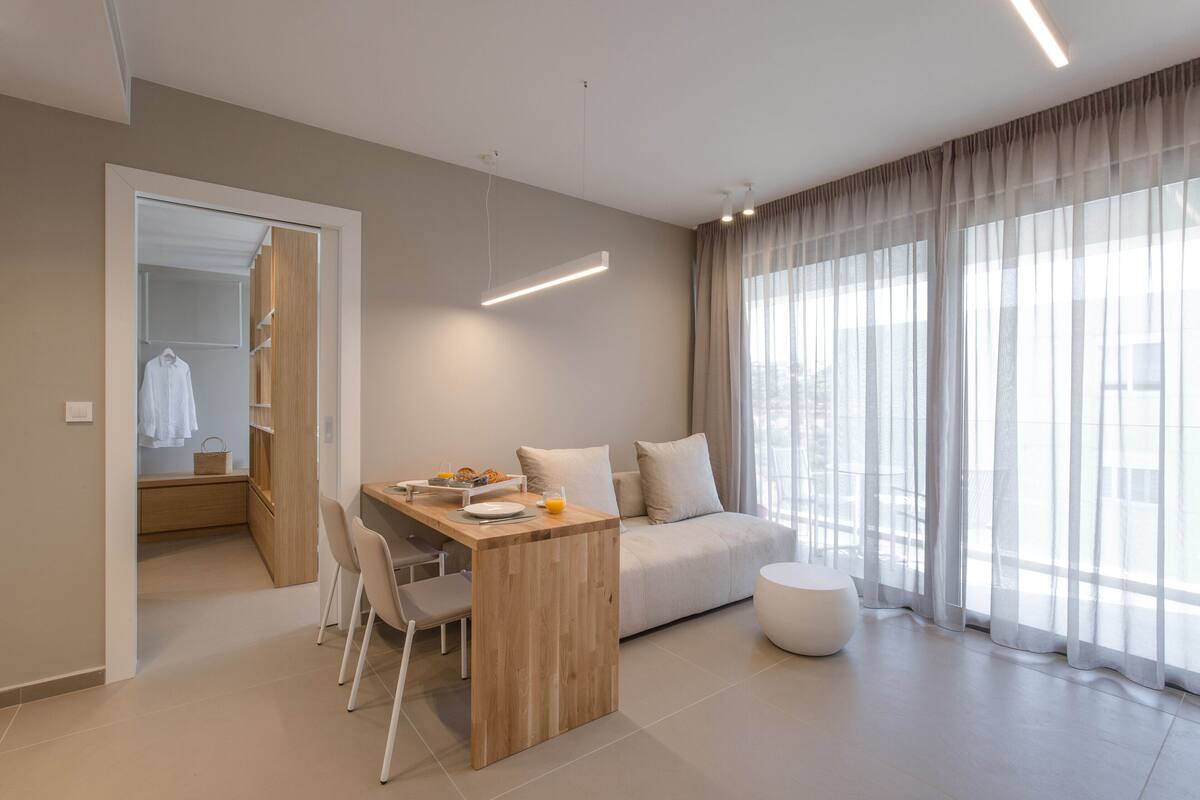 Una sala de estar moderna con una mesa de comedor de madera, un sofá y un gran ventanal con cortinas translúcidas.