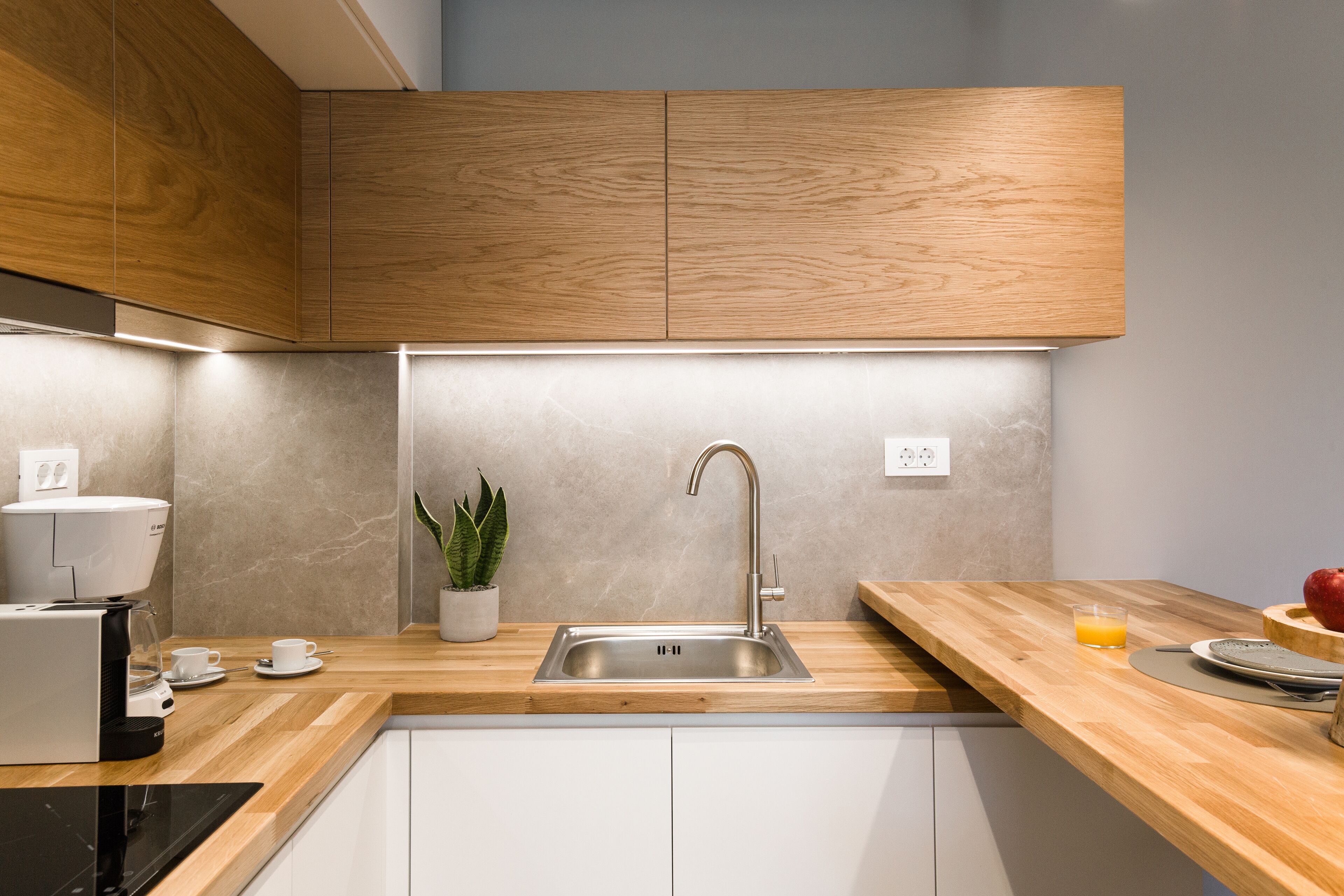 A modern kitchen with wooden cabinets, a stainless steel sink, a coffee machine, and a potted plant.