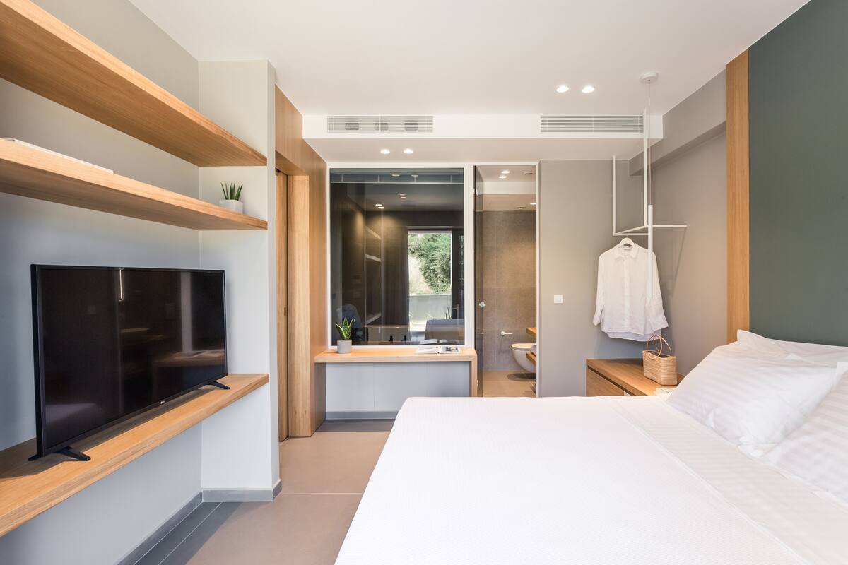 Una habitación de hotel moderna con televisión de pantalla plana, una cama, una mesita de noche, una toalla colgada y vistas al baño.