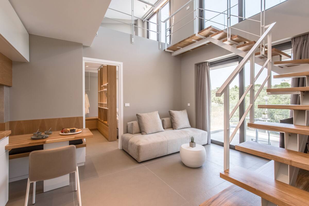 Un interior moderno con una escalera tipo loft, un sofá, una zona de comedor y vistas amplias a través de grandes ventanales.