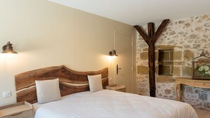 A bedroom with a wooden headboard, a bed with white bedding, a nightstand, a wall-mounted lamp, and a stone wall with a small window.