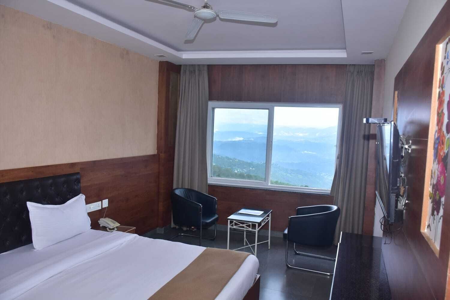Une chambre d’hôtel avec un lit, un bureau, deux chaises, un ventilateur de plafond et une grande fenêtre donnant sur un beau paysage.