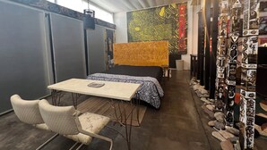 A cozy room with a bed, a table, and a chair. There is a colorful tapestry on the wall and a decorative pillar with various designs.
