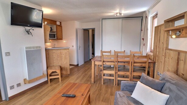A living room with a sofa, wooden dining table, chairs, a TV mounted on the wall, and a kitchen area with wooden cabinets and a microwave. - Comfortable 2 Rooms + Cabin, Pets Allowed, Ski-in/Ski-out (La Tania)