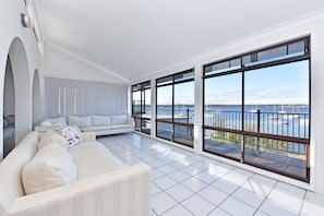 A spacious living area with a large window overlooking a body of water, a white sofa, and a tiled floor.