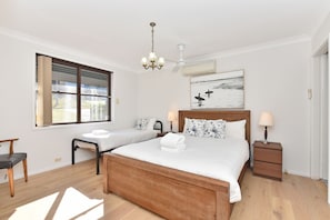 A hotel room with a large bed, two smaller beds, a desk, a chair, a window with blinds, a chandelier, an air conditioning unit, a bedside table with a lamp, and a wall-mounted artwork.