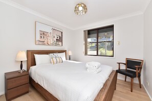 A neatly arranged bedroom with a bed, two bedside tables, a chair, a window with a view, and a wall-mounted light fixture. - Sunshine Shores Absolute Waterfront (Sunshine)