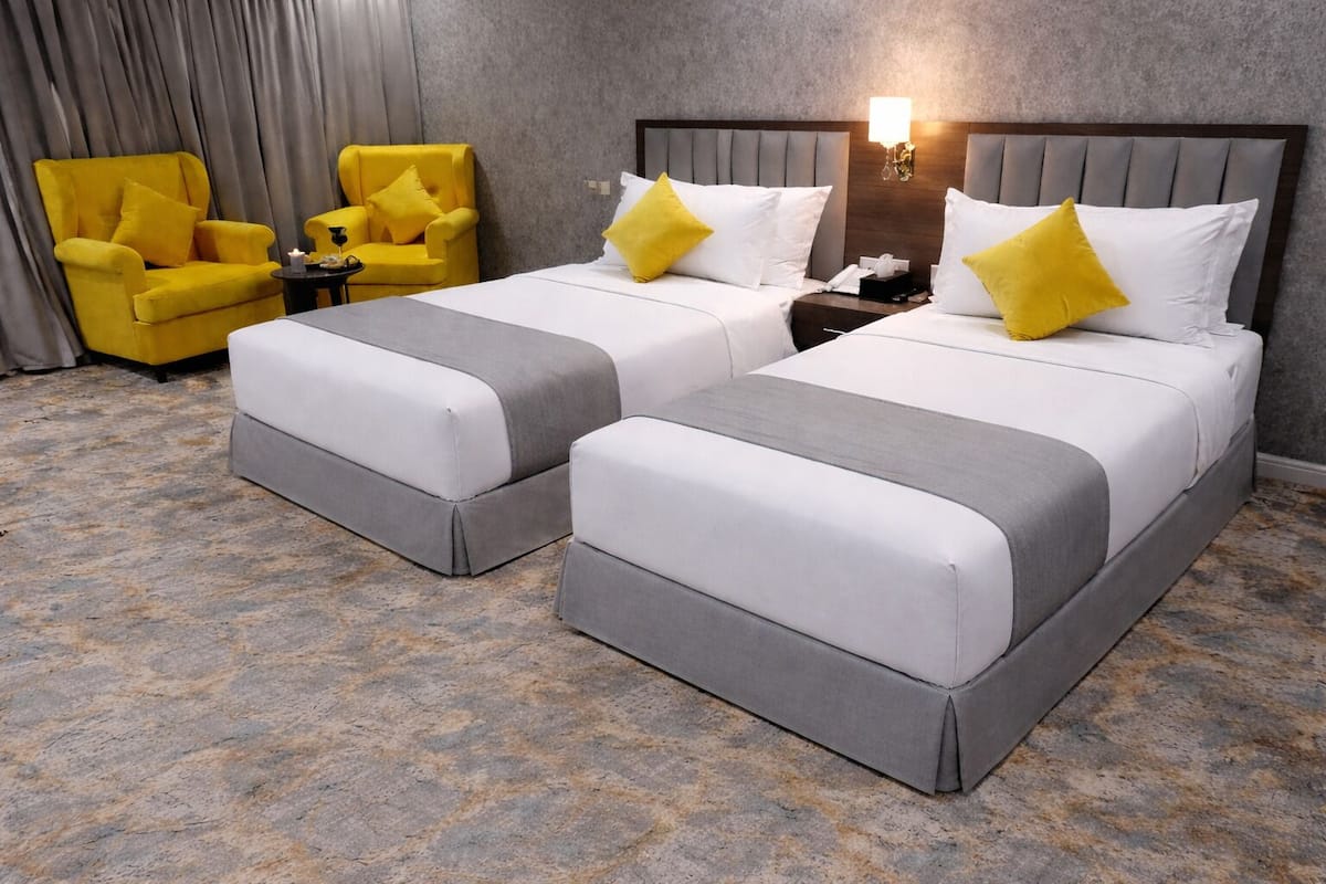 A hotel room with two beds, a nightstand, a lamp, and two yellow armchairs.