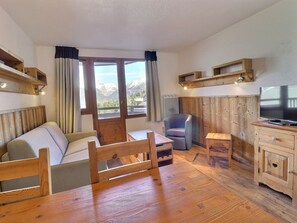 A cozy room with a wooden bed, a small table, a chair, a TV, and a view of mountains through large windows.