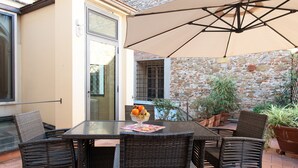 Een terras buiten met een glazen tafel, rieten stoelen, een grote parasol en potplanten.
