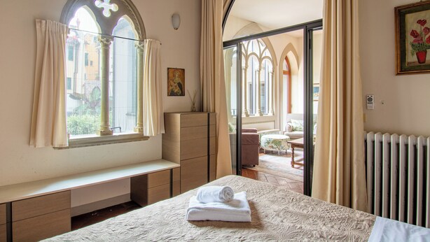 Een hotelkamer met een bed, een bankje, een raam met gordijnen, een radiator en een schilderij aan de muur.
