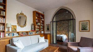 Een gezellige woonkamer met een bank, fauteuil en salontafel. Boekenkasten vol boeken en een grote spiegel zijn zichtbaar. Achter een glazen deur is een bed te zien.