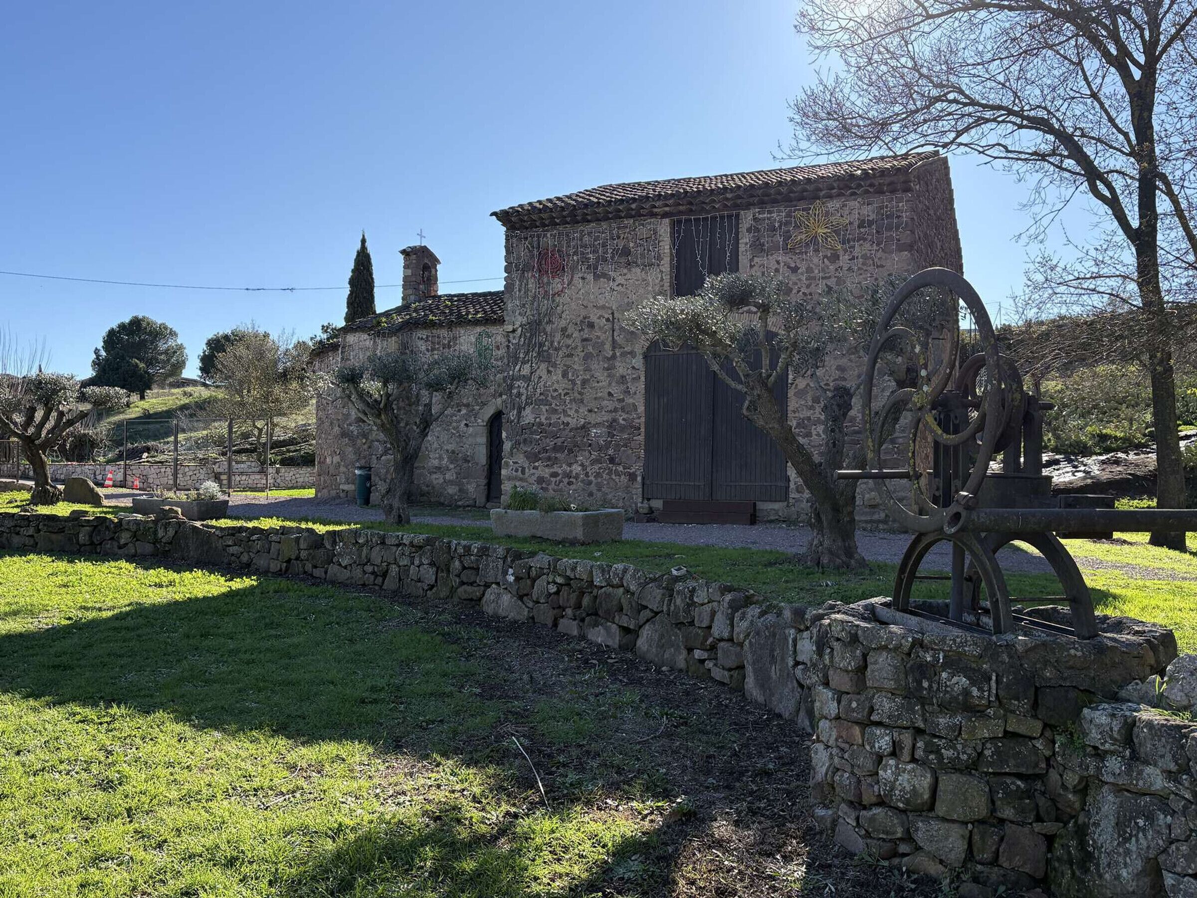 Un antiguo edificio de piedra con un campanario, rodeado por un muro de piedra y vegetación.