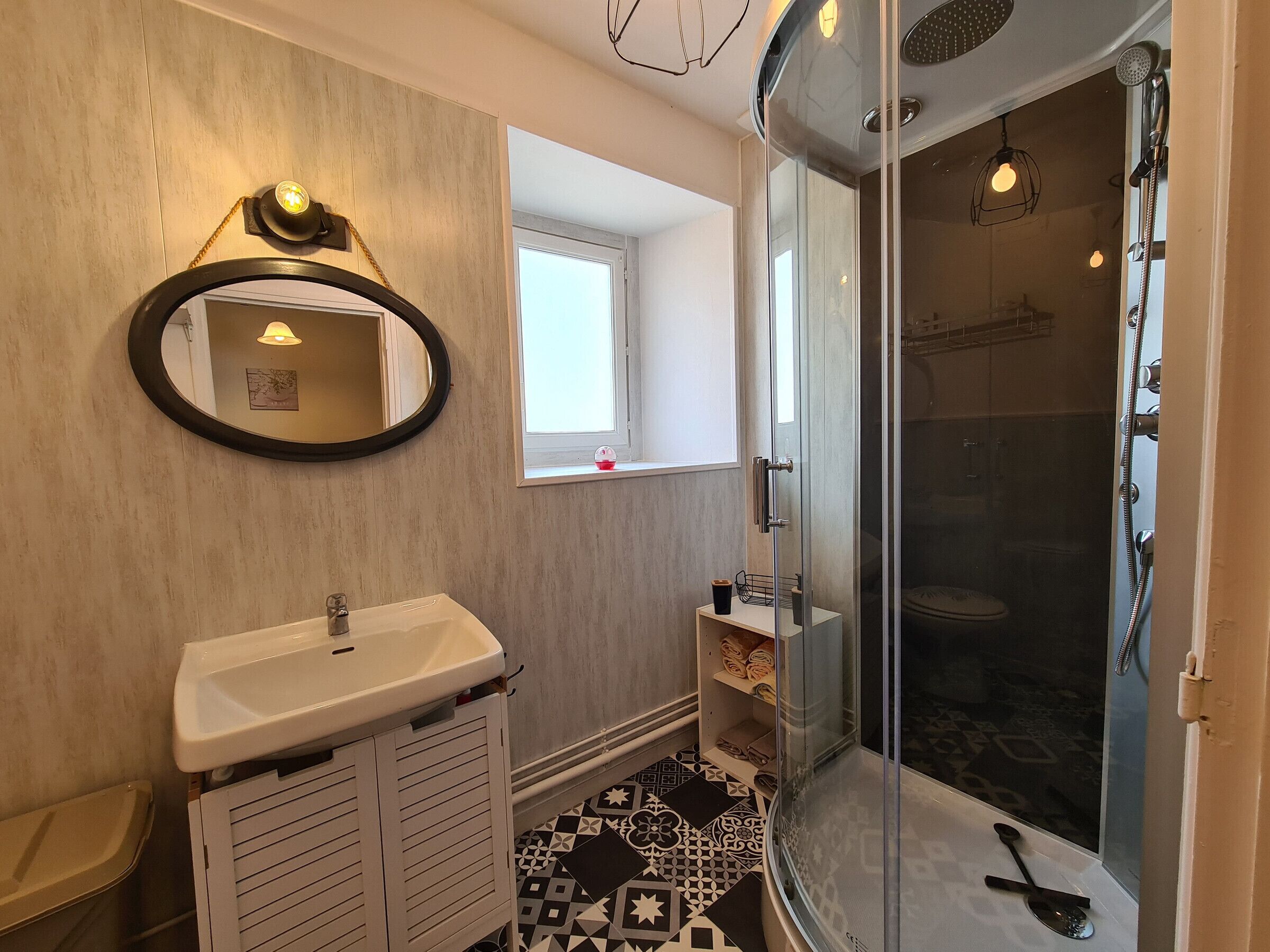Een badkamer met een wit wastafelmeubel, een spiegel, een douche met glazen deur en een raam dat voor natuurlijk licht zorgt.