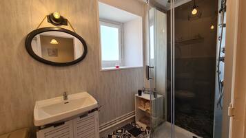 Een badkamer met een wit wastafelmeubel, een spiegel, een douche met glazen deur en een raam dat voor natuurlijk licht zorgt.