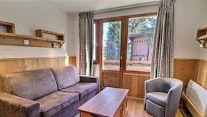 Een gezellige woonkamer met een houten bank, een fauteuil, een houten salontafel met wijnflessen, en een groot raam met gordijnen.